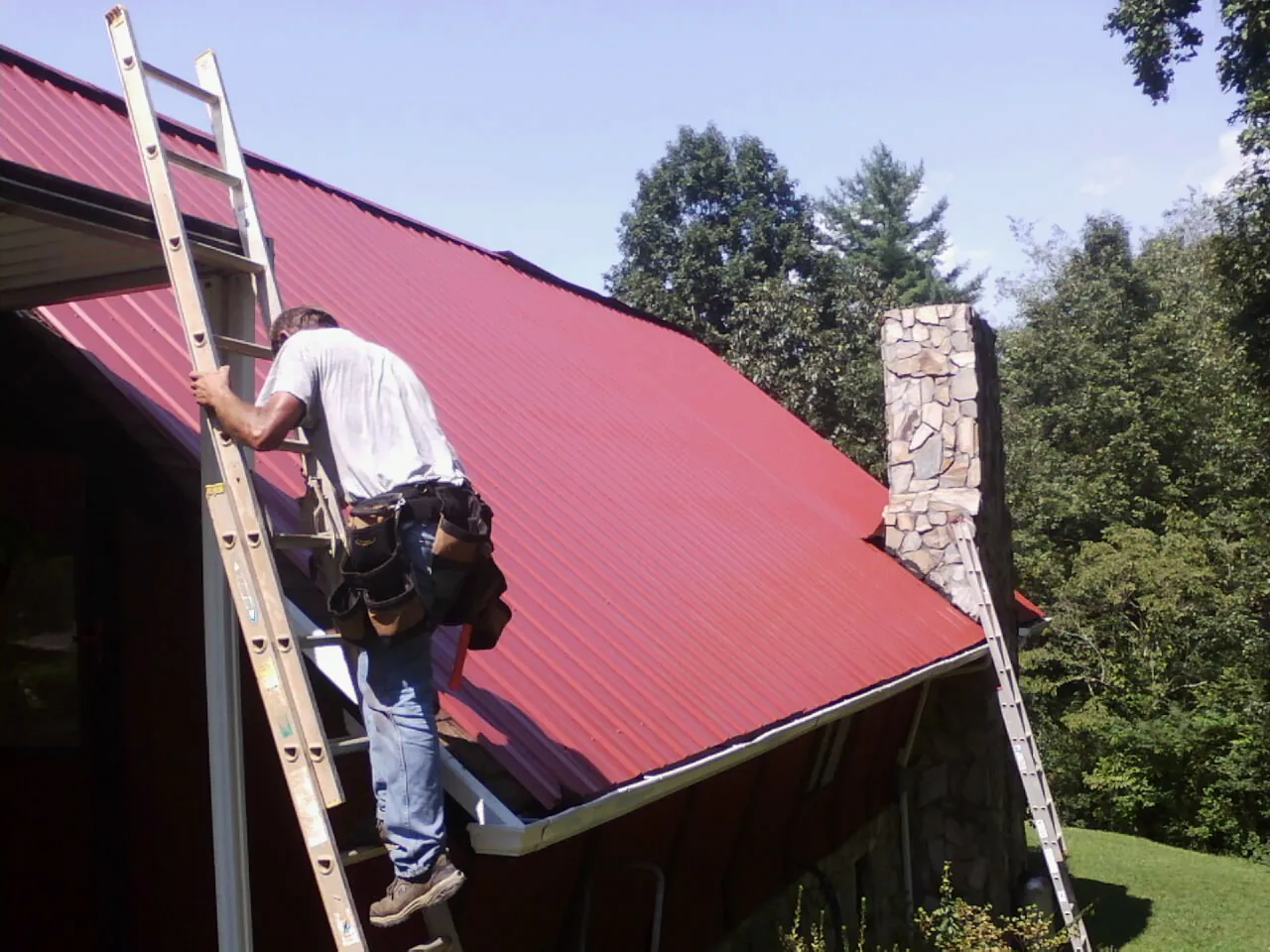 Roofer on a ladder beside a red metal roof and stone chimney — Soffit & Fascia Repair by Roofing Brothers in Gorham