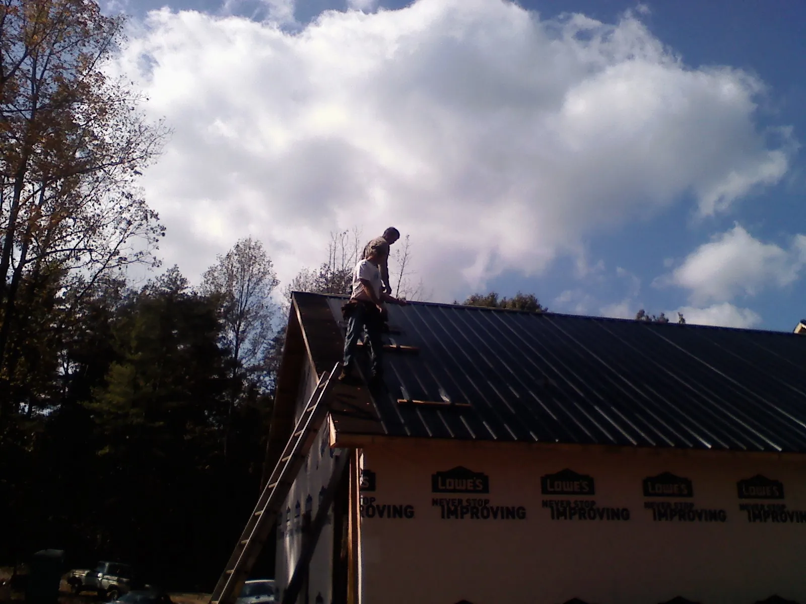 Roofer fastening dark metal panels high against a cloudy sky, from a Roof Inspection job in Gorham